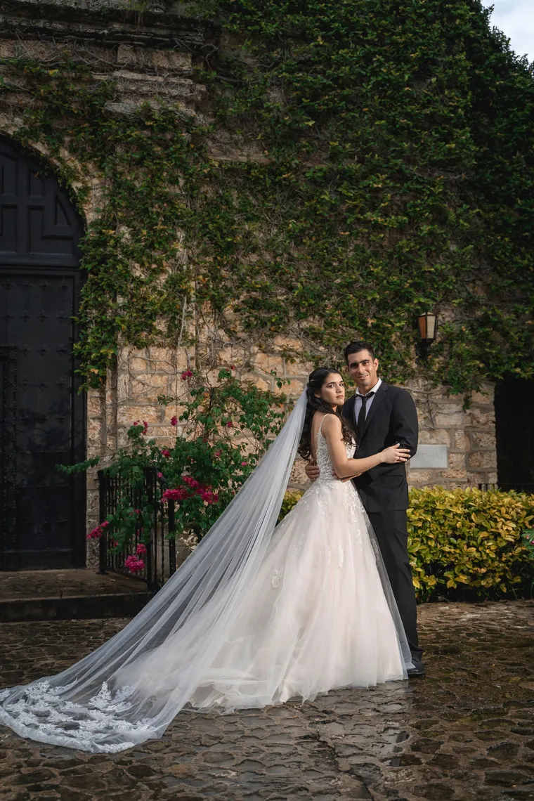 Novios abrazados junto a un muro de hiedra, el velo extendido sobre los adoquines, de Photoluv Studios en Miami