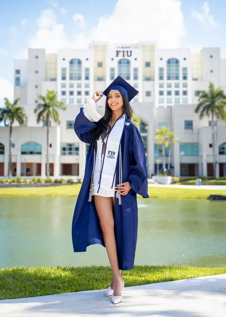 Graduada con toga azul abierta sobre vestido blanco junto a un lago y edificio blanco, de Photoluv Studios en Miami