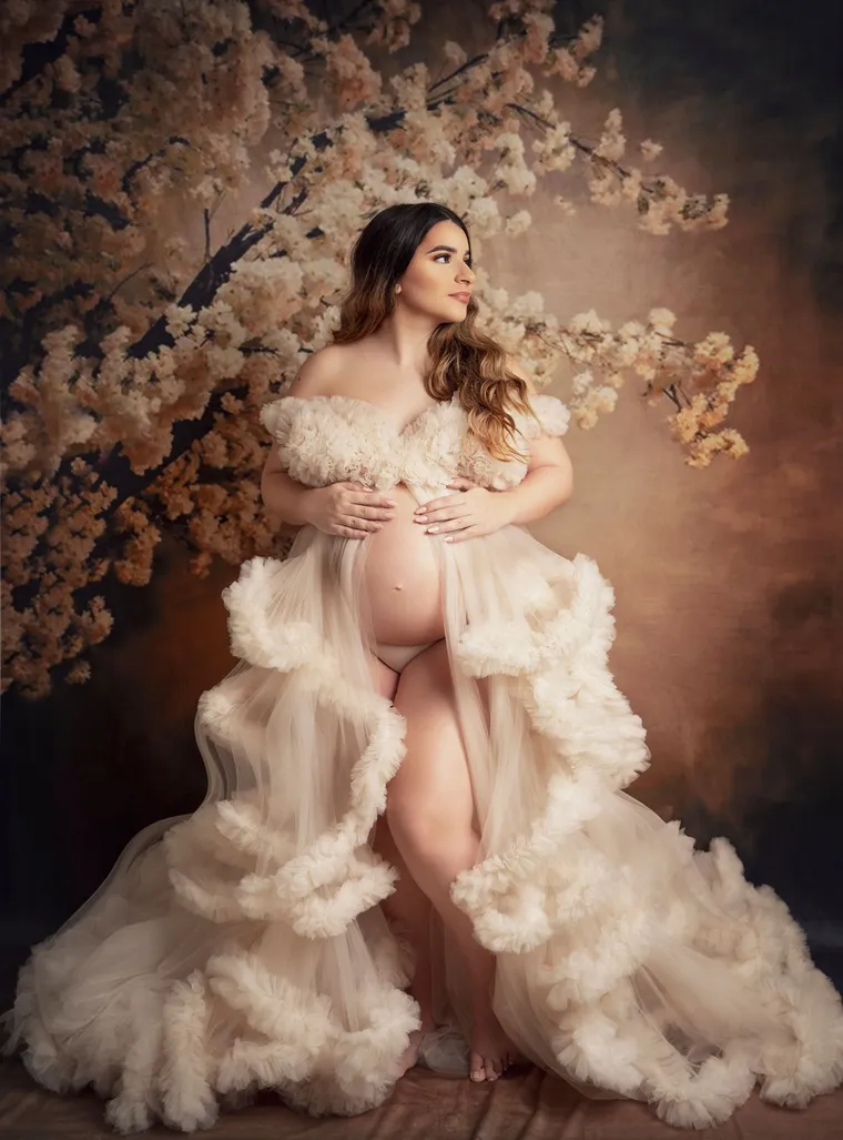 Mujer embarazada con bata de tul crema ante ramas de flores blancas, luz cálida, de Photoluv Studios en Miami